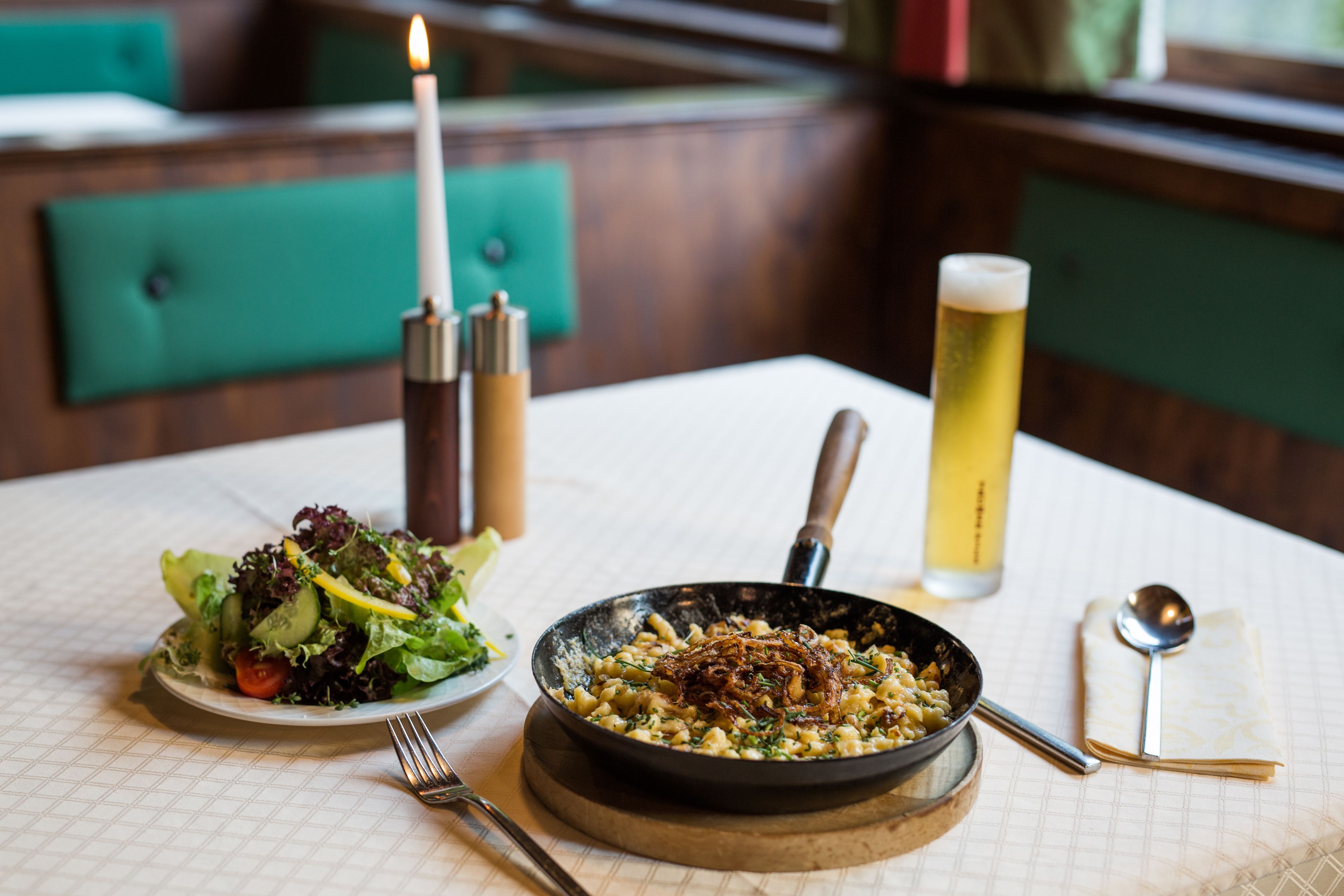 A dining table with a plate of food, a bowl of salad, a candle, a spoon, and a glass of beer.