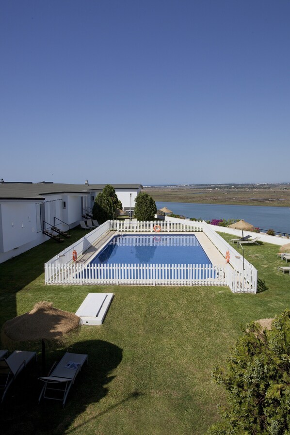 A rectangular swimming pool surrounded by a white picket fence, with a grassy lawn and lounge chairs.