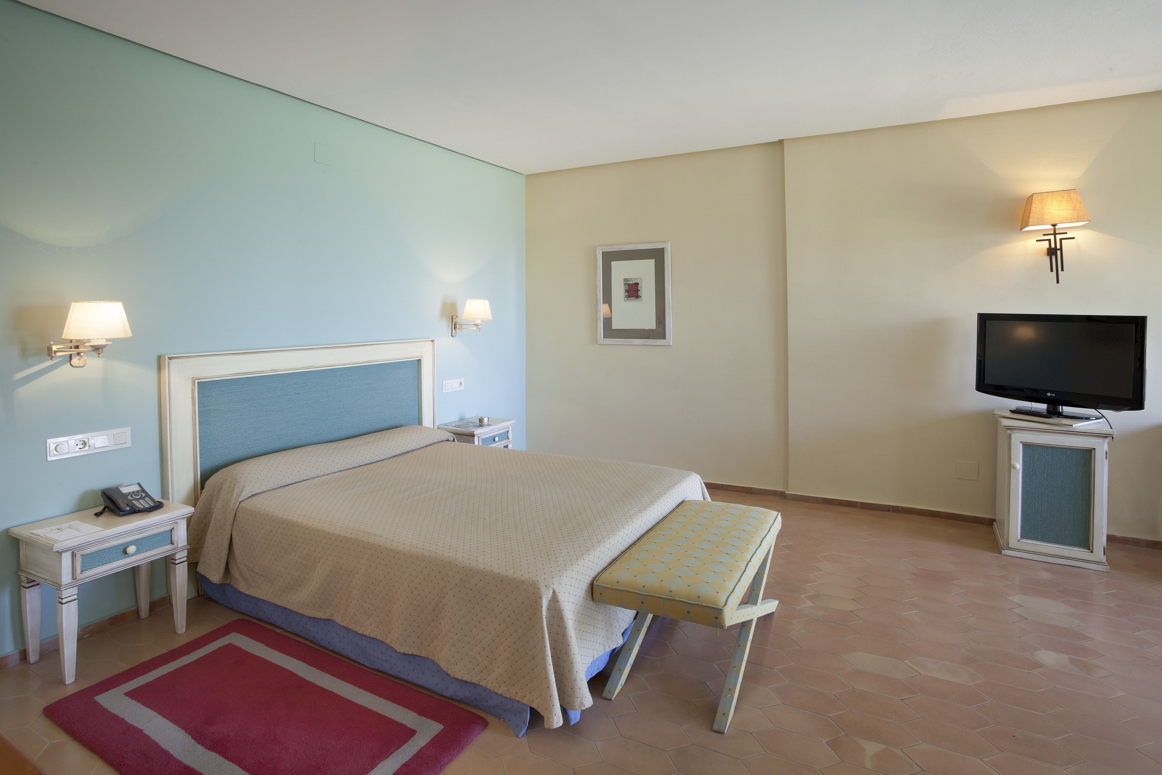 A hotel room with a bed, bedside tables, a TV, a small table, and a red rug.