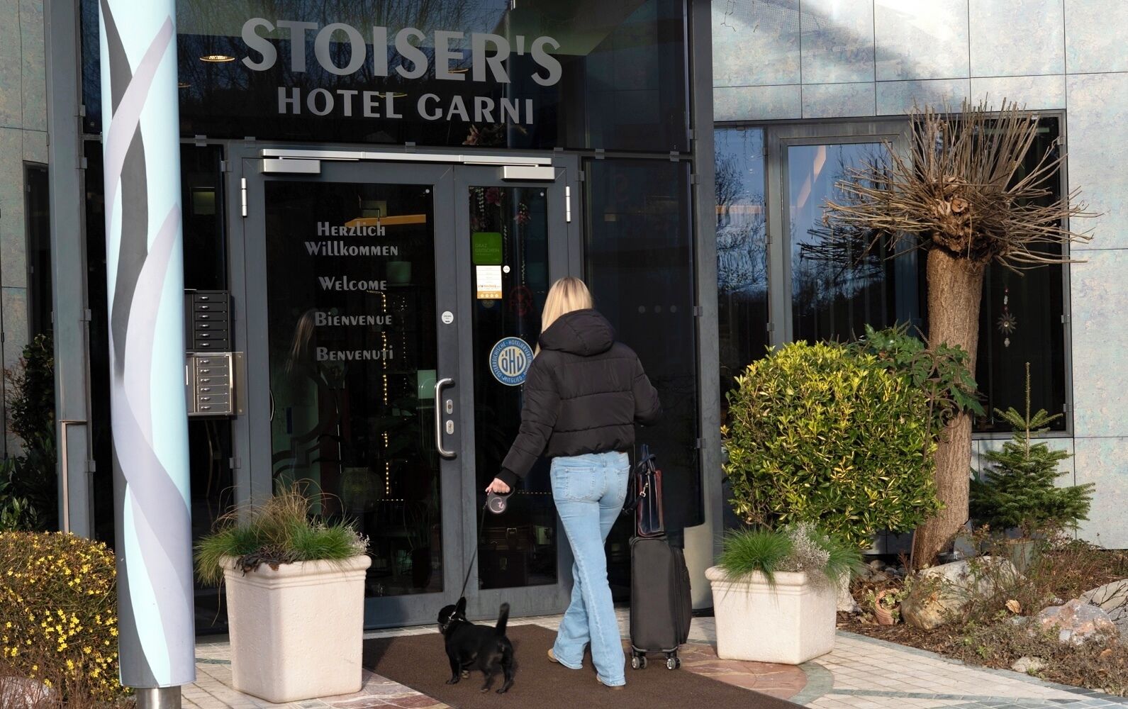 The entrance to Stoiser's Hotel Carni with a modern glass facade, a welcome sign, and potted plants.