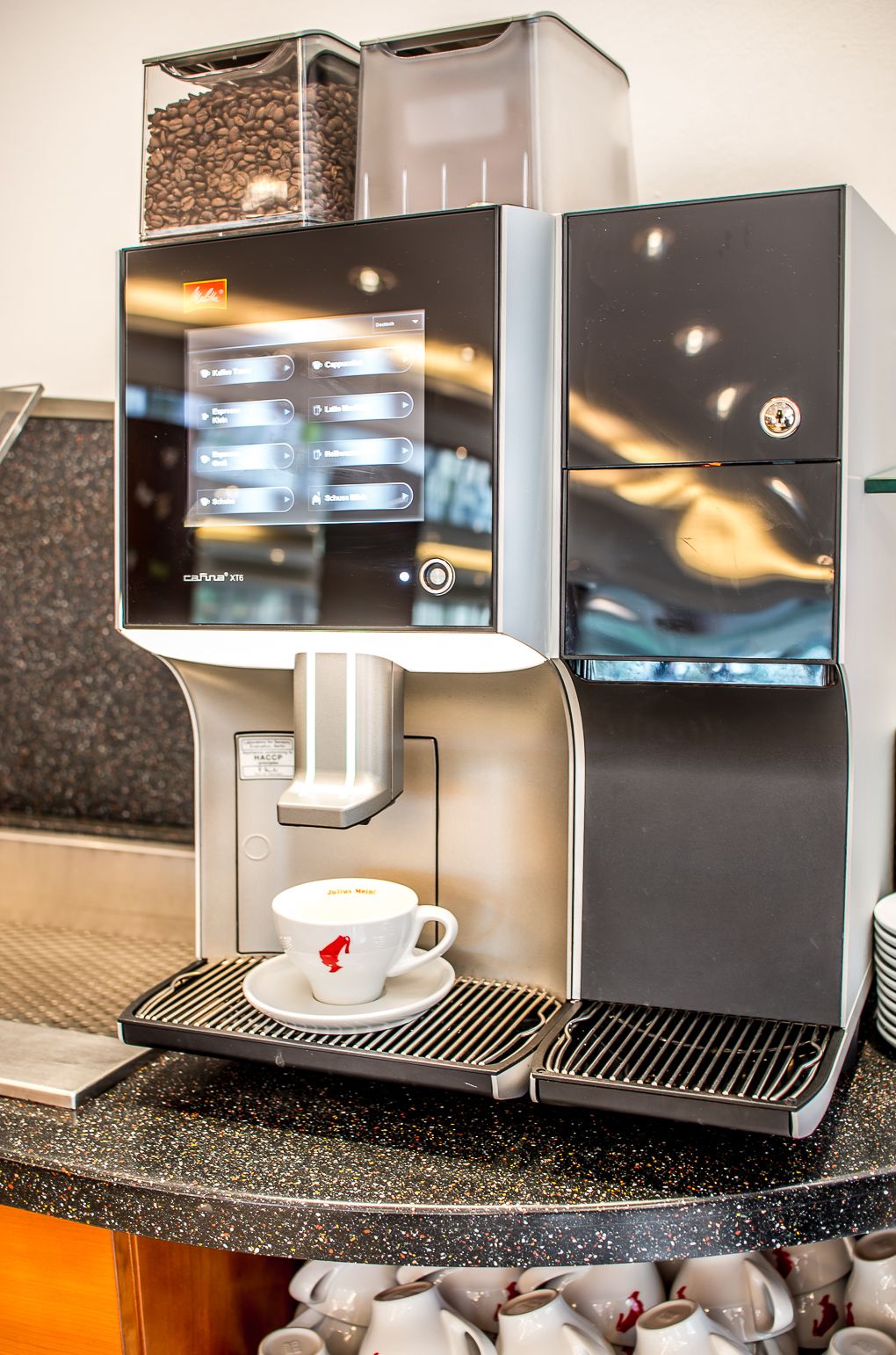 Una cafetera con pantalla táctil, una taza de café y una pila de vasos desechables.