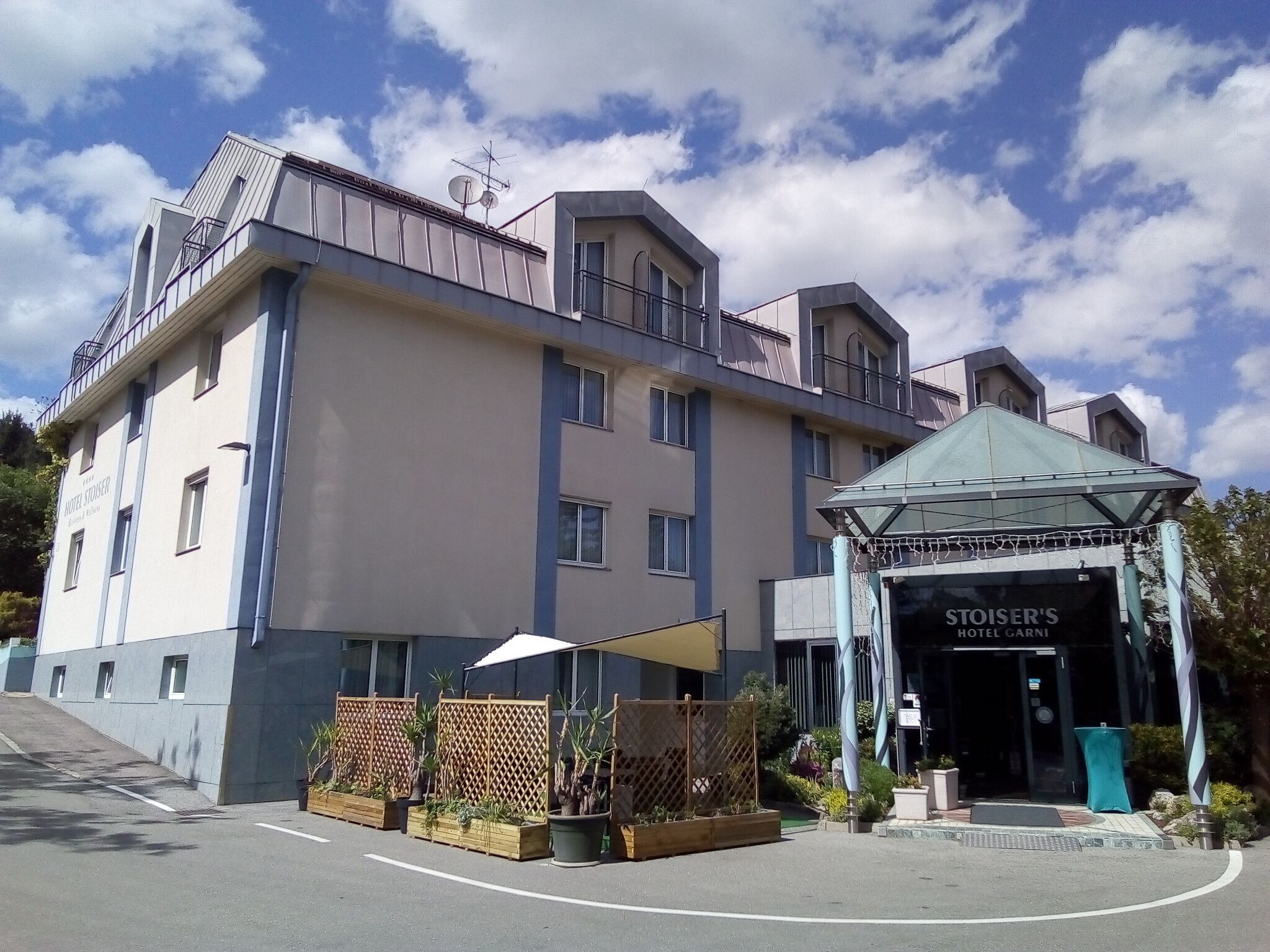 Um hotel com design moderno, apresentando uma entrada imponente com uma cobertura de vidro e uma placa com a inscrição "Stoiser's Hotel Carnig". O edifício possui vários andares, varandas e uma área externa bem cuidada, com plantas e um espaço para sentar.