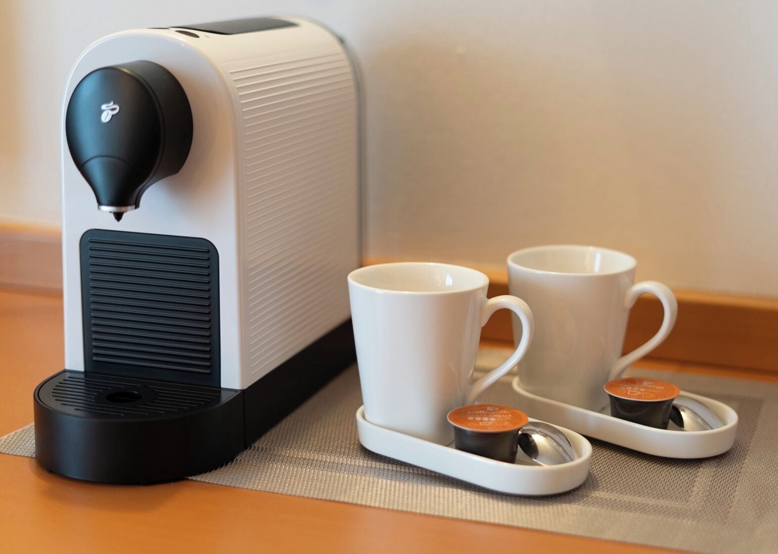 En kaffemaskine med to hvide kopper og underkopper på en vævet bordskåner.