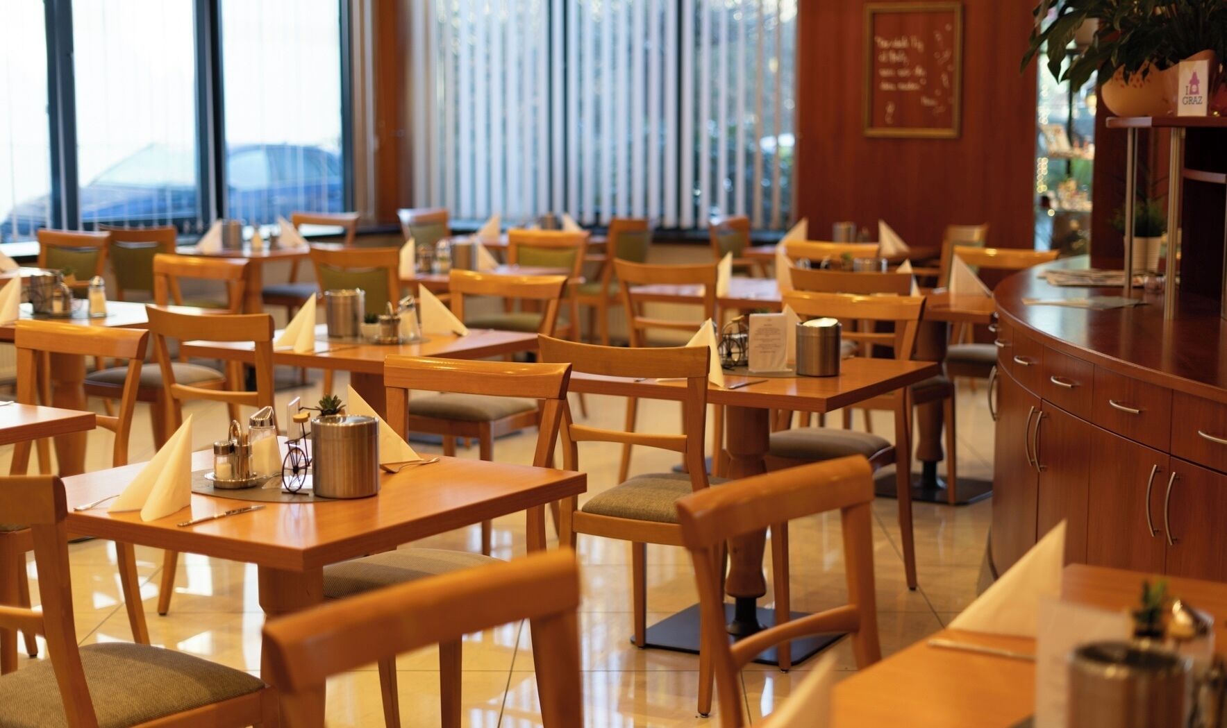 A restaurant with wooden tables and chairs, a bar area, and a view of the city through large windows.