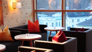 A modern lounge area with sofas and round tables, large windows with a view of buildings, and a wall-mounted light fixture.