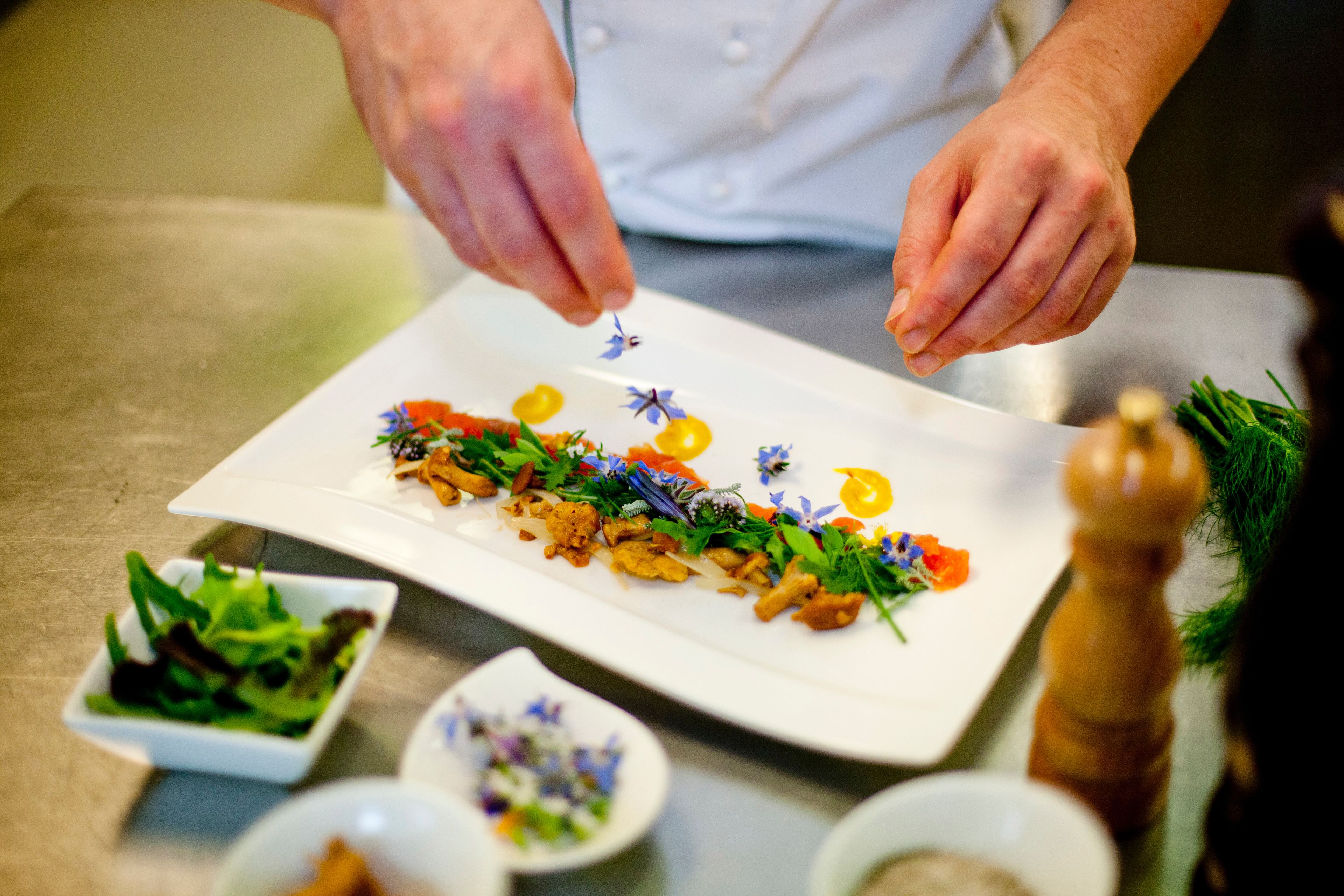 A chef plating a dish with various garnishes and a small bowl of greens.