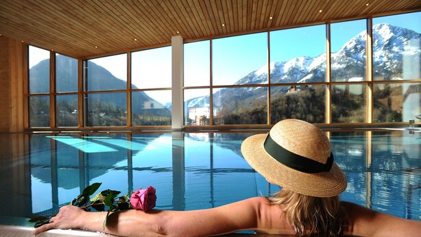 A person relaxing by a pool with a mountain view through large windows.