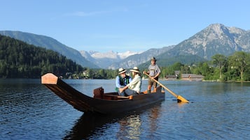 A wooden boat on calm water with people rowing, surrounded by mountains and houses in the background.
