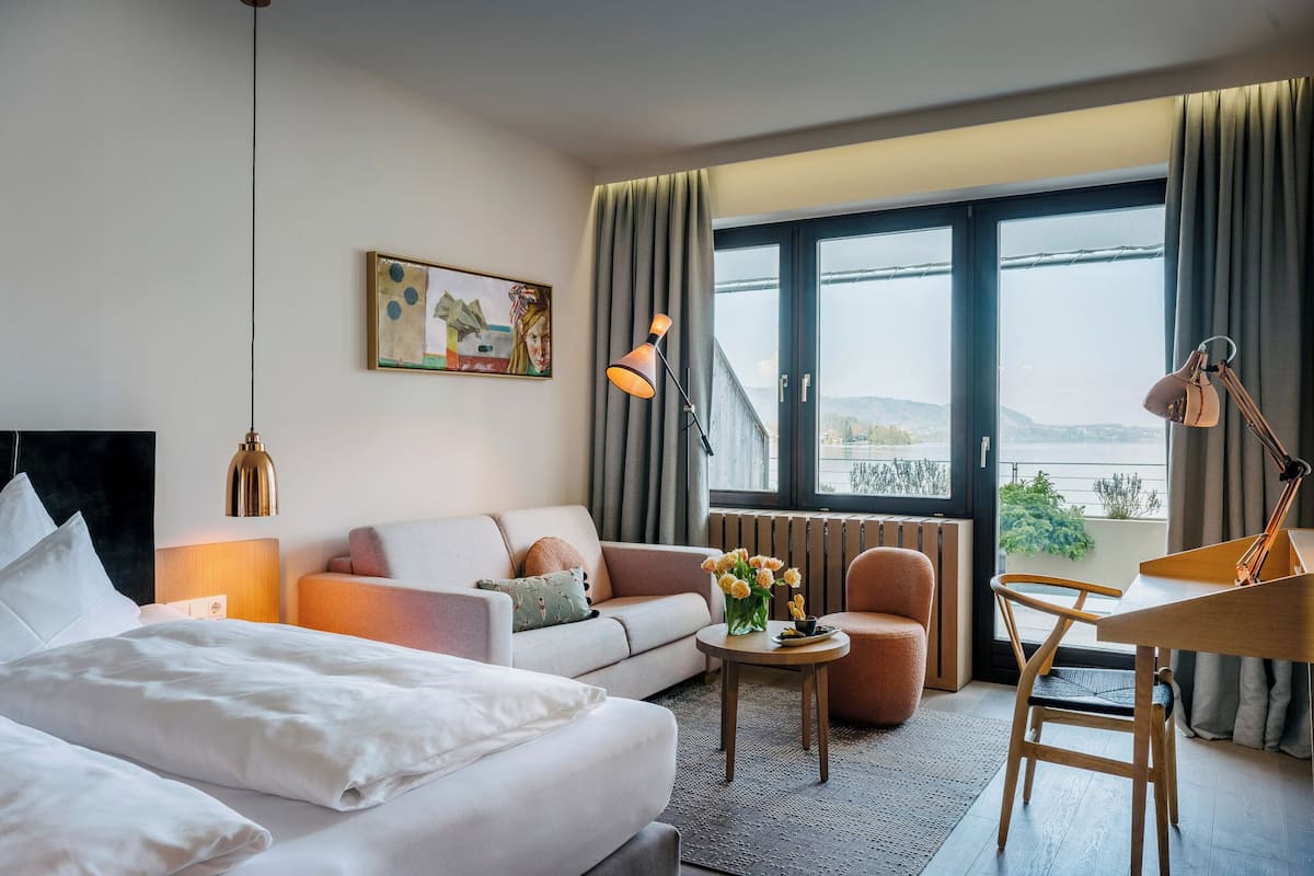 A hotel room with a bed, a sofa, a dining table, a chair, a lamp, a vase with flowers, a painting on the wall, and a large window with curtains.