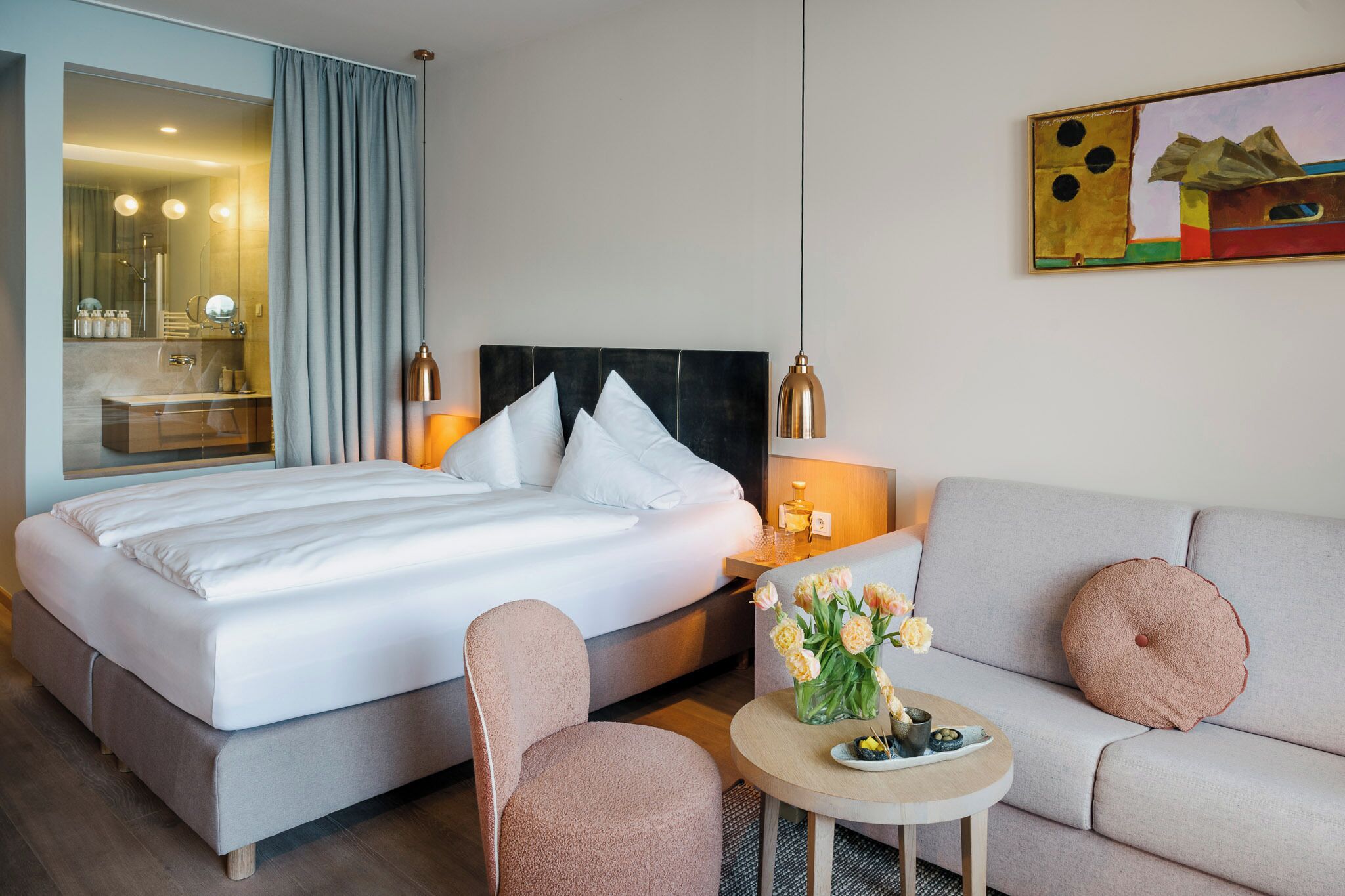 Una habitación de hotel con cama, sofá, una mesita con flores, espejo y un cuadro en la pared.
