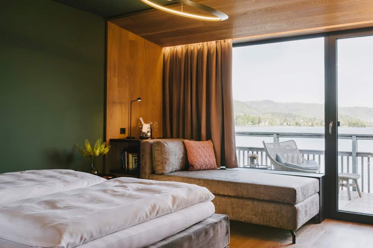 A modern bedroom with a large bed, a sofa, and a balcony with a view of the hills.