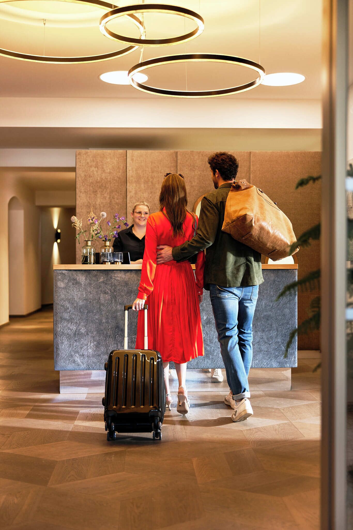 A reception desk with a modern design, a person in a red dress with a suitcase, and another person carrying a large bag.