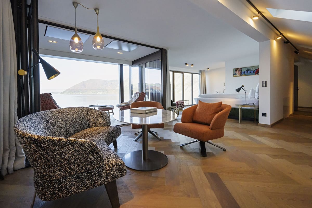 A modern hotel room with a large window offering a view of the sea, a round dining table, and contemporary furniture.