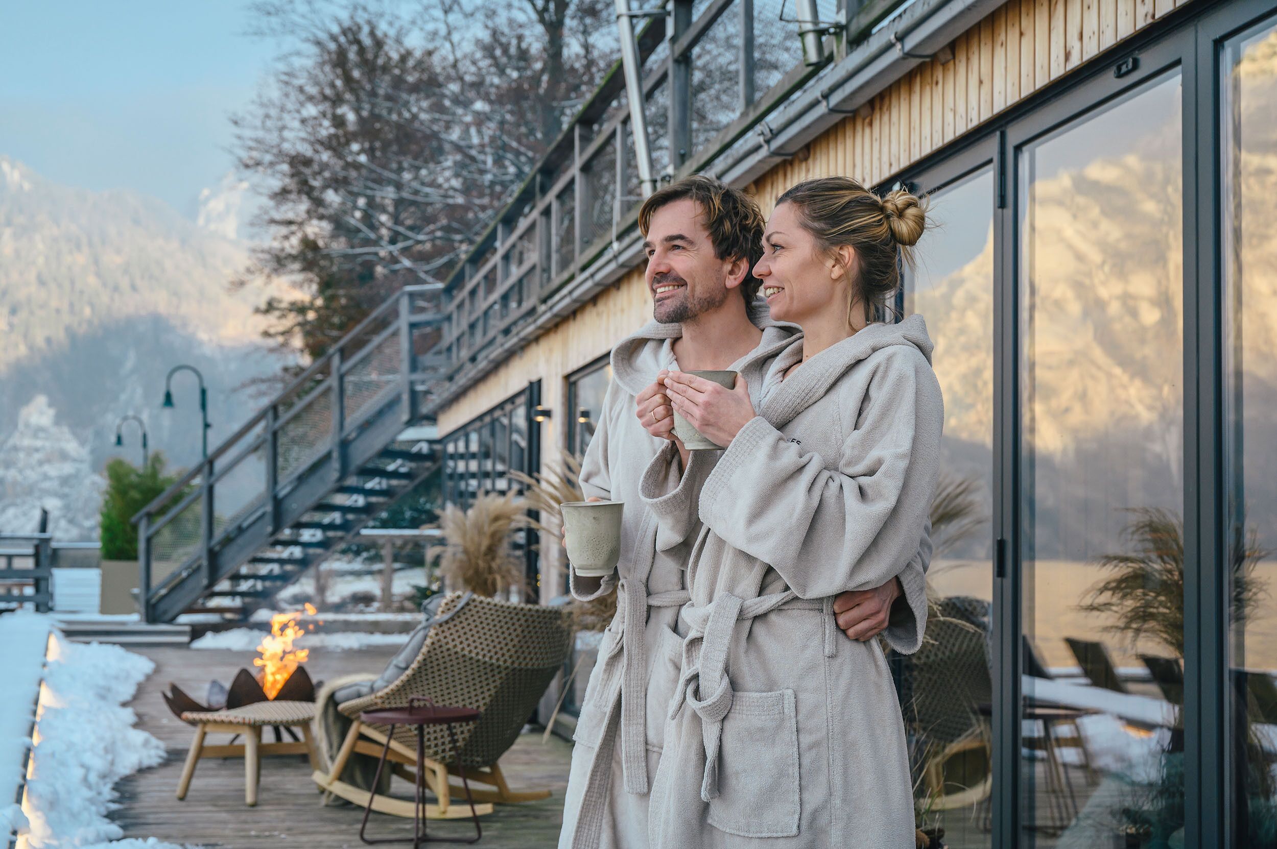 Pariskunta seisoo puukannella kylpytakeissaan nuotion ääressä. He katselevat lumista maisemaa ja suurilla ikkunoilla varustettua rakennusta.