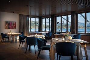 A restaurant with tables and chairs, large windows with a view of a body of water and a bridge, and a ceiling with a wooden pattern.