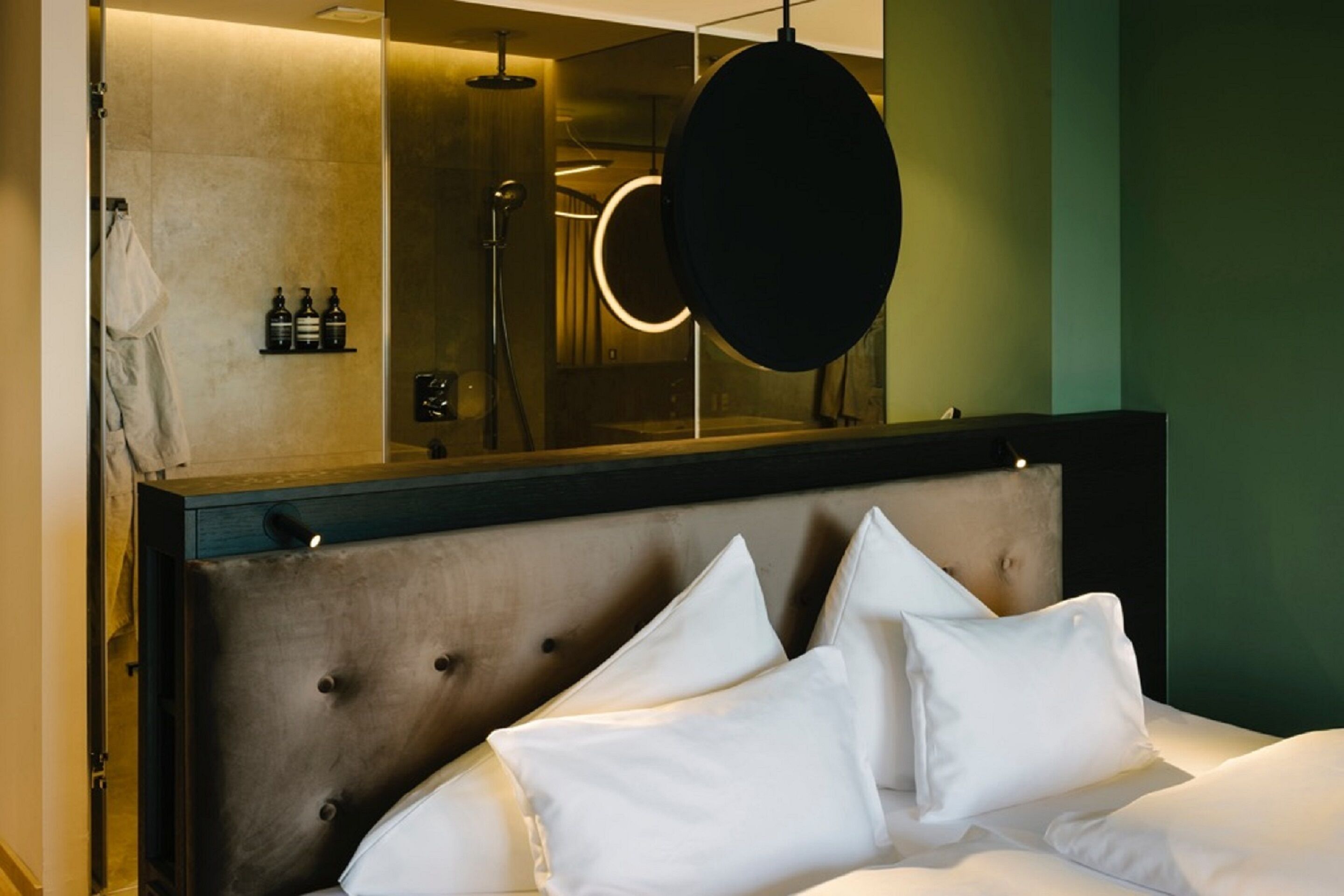 A modern hotel room with a large bed, a headboard with tufting, a round mirror, and a wall-mounted shelf with toiletries.