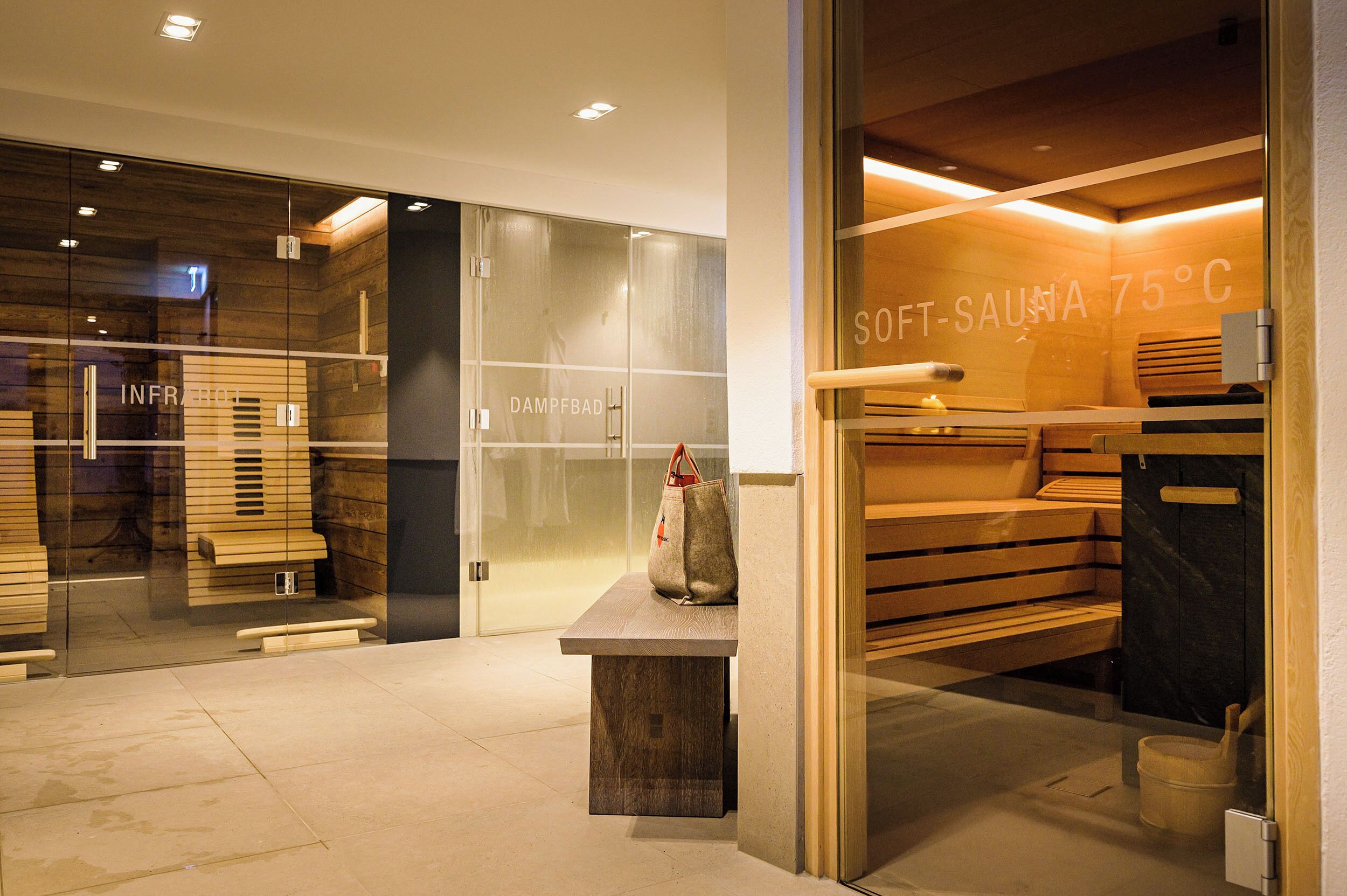 Un spa moderno con paredes de vidrio, bancos de madera y un cartel que indica una sauna suave a 75°C.