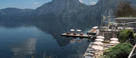 A lakeside dock with lounge chairs and umbrellas, a wooden deck, and mountains in the background.