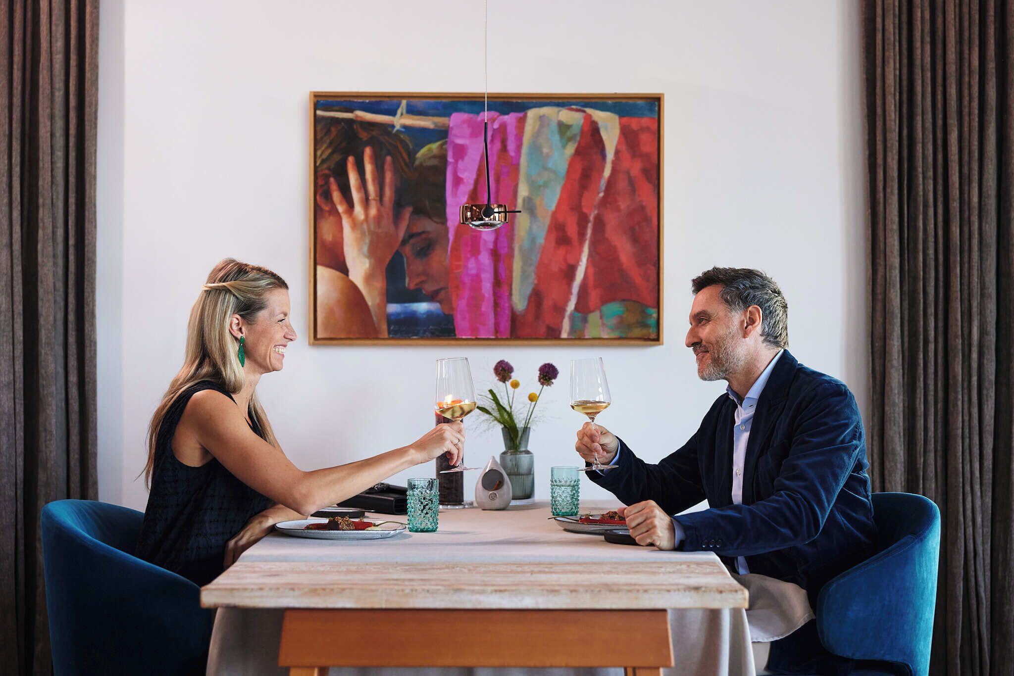 Una mesa de comedor con dos personas brindando con copas de vino, un florero con flores y un cuadro en la pared.