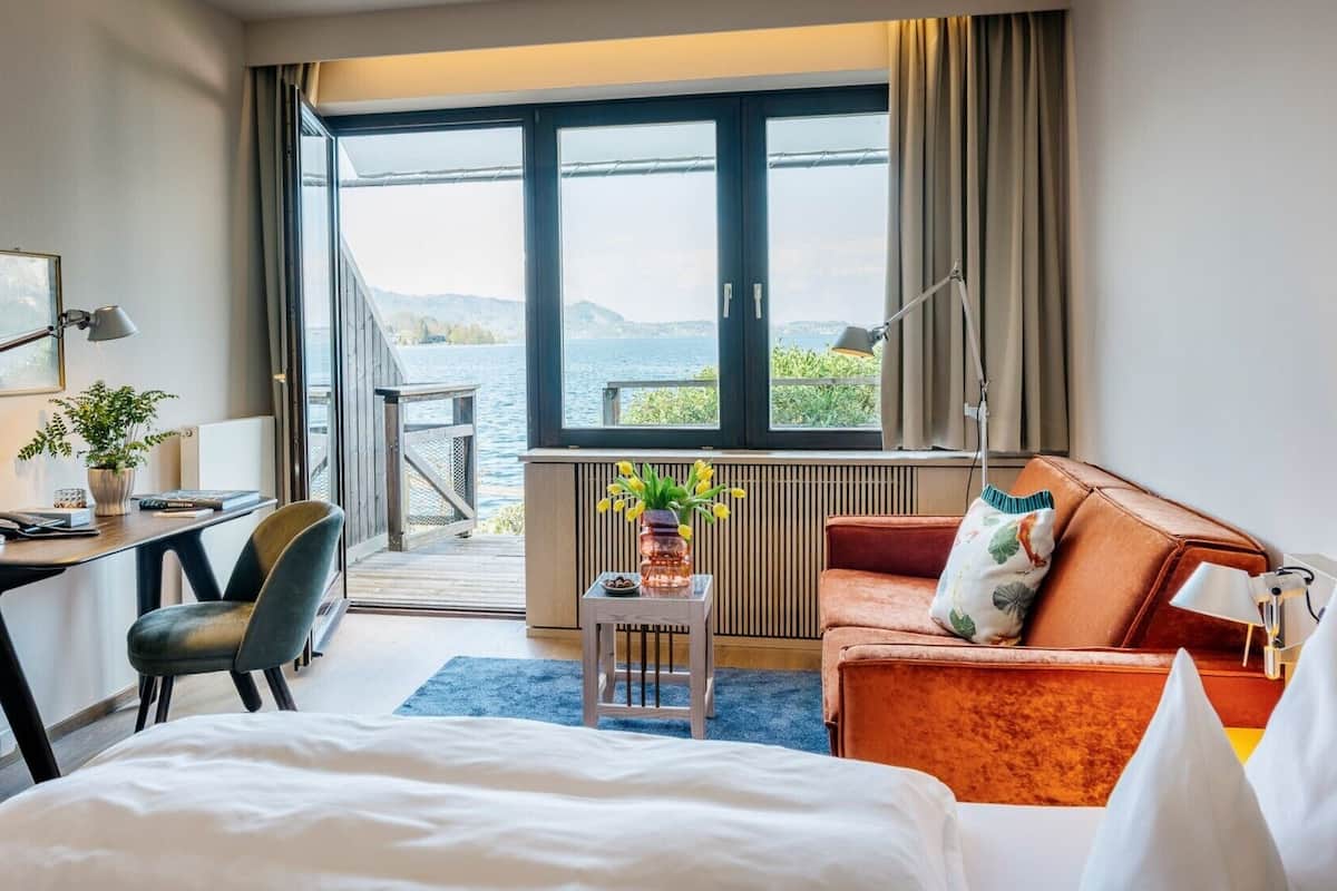 A hotel room with a bed, a desk, a chair, a sofa, a view of the water, and a window.