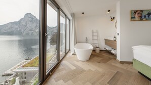 A modern bathroom with a freestanding bathtub, a wooden floor, and a large window overlooking a body of water and mountains. - Das Traunsee (Traunkirchen)