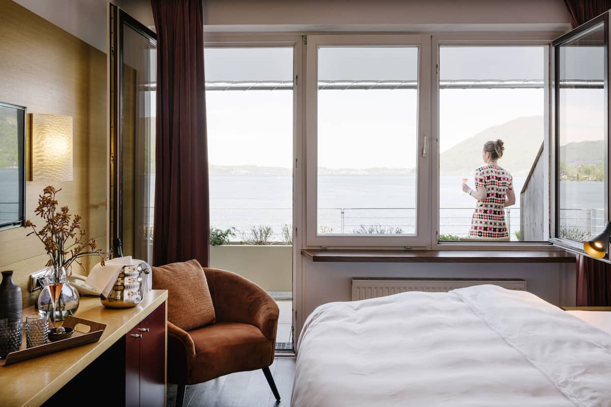 A hotel room with a large window, a bed, a desk with a vase, and a chair.