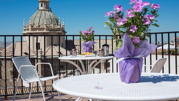 Une terrasse avec une table blanche, des chaises et un vase de fleurs. Au fond, on aperçoit un bâtiment doté d’un dôme et d’une clôture.