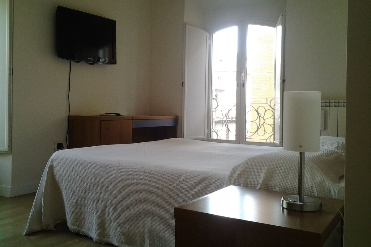 A hotel room with a bed, a desk, a lamp, a TV mounted on the wall, and a balcony with a wrought iron gate.