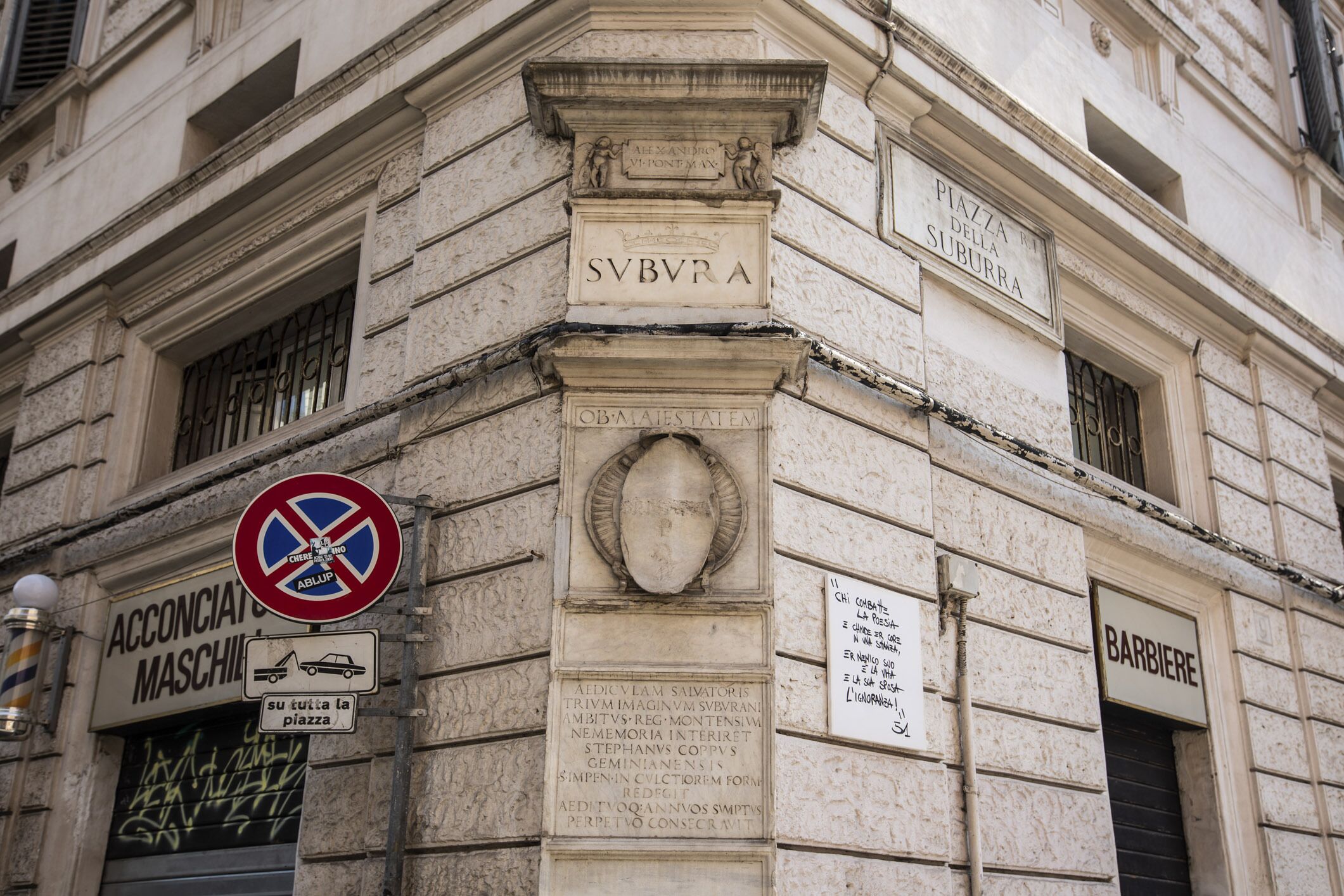 A building facade with signs reading "SVBVRA," "Piazza della Suburra," and "Barbiere."