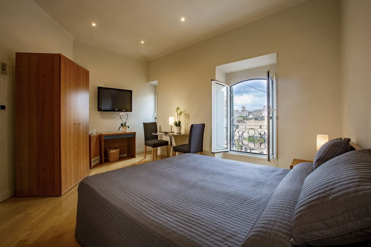 Une chambre avec un grand lit, une armoire en bois, un bureau avec une chaise, un téléviseur fixé au mur et un balcon avec vue sur la ville.