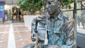 Una estatua de bronce que representa a un hombre sentado con una guitarra, ubicada en una plaza al aire libre con suelo de baldosas y vegetación de fondo.
