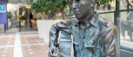Una estatua de bronce que representa a un hombre sentado con una guitarra, ubicada en una plaza al aire libre con suelo de baldosas y vegetación de fondo.