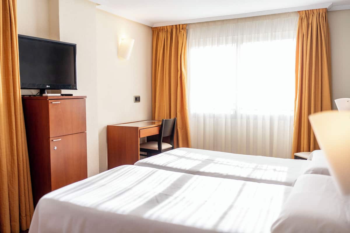 A hotel room with two beds, a desk with a chair, a television on a cabinet, and curtains on the windows.