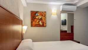 Habitación de hotel con cama, un cuadro en la pared, lámpara, equipo de aire acondicionado y un espejo que refleja un televisor y un baño.