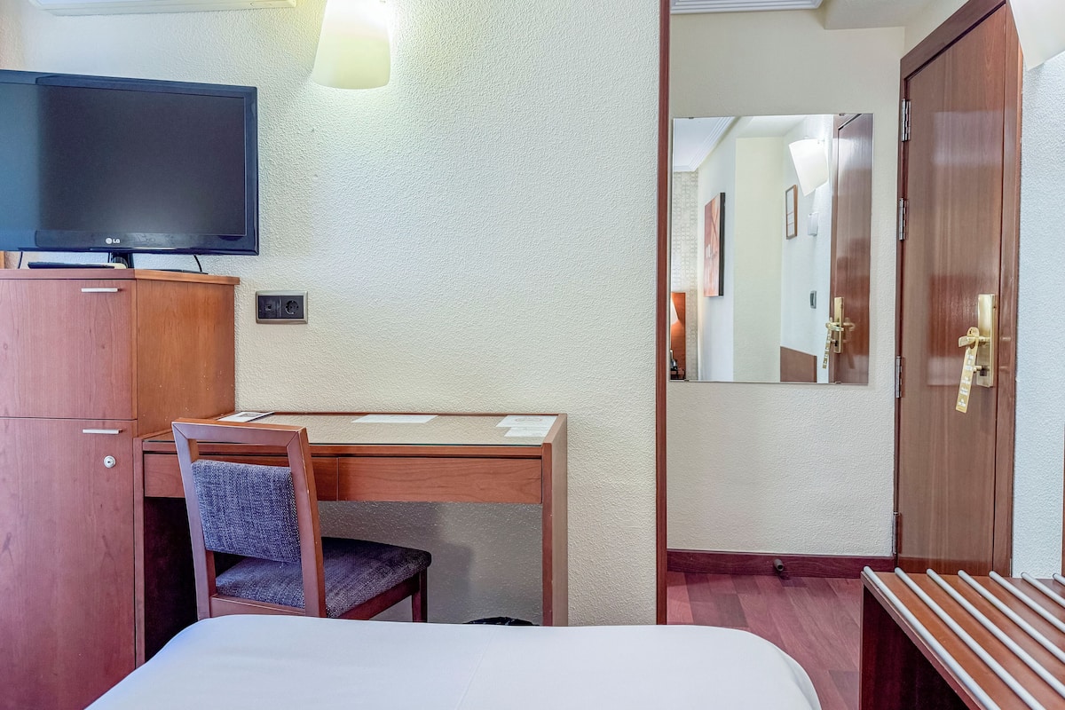 A hotel room with a bed, a desk with a chair, a television, and a wardrobe.