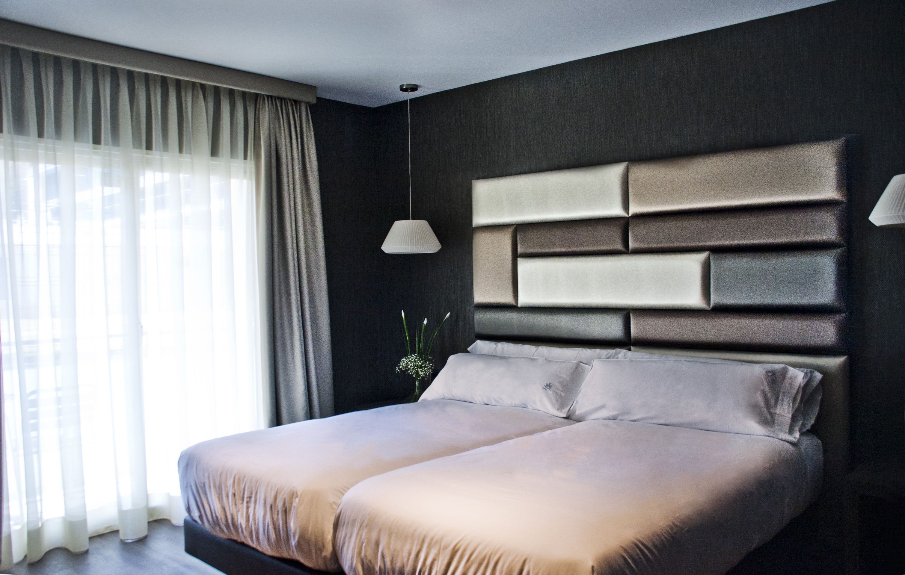 Quarto moderno com uma cama grande, parede escura e janela com cortinas leves.