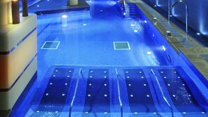 Una piscina interior moderna con suelo iluminado en azul, barandillas de vidrio y una escalera que sube a niveles superiores.