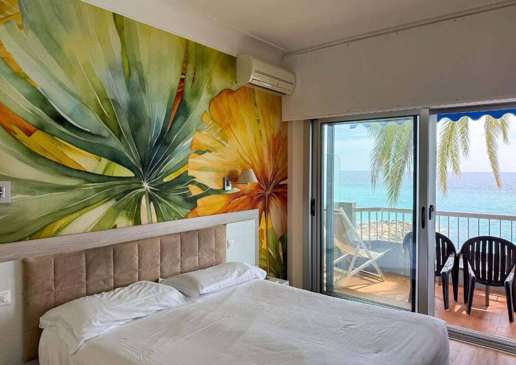 A hotel room with a bed, a large tropical mural, and a balcony with a view of the sea and palm trees.