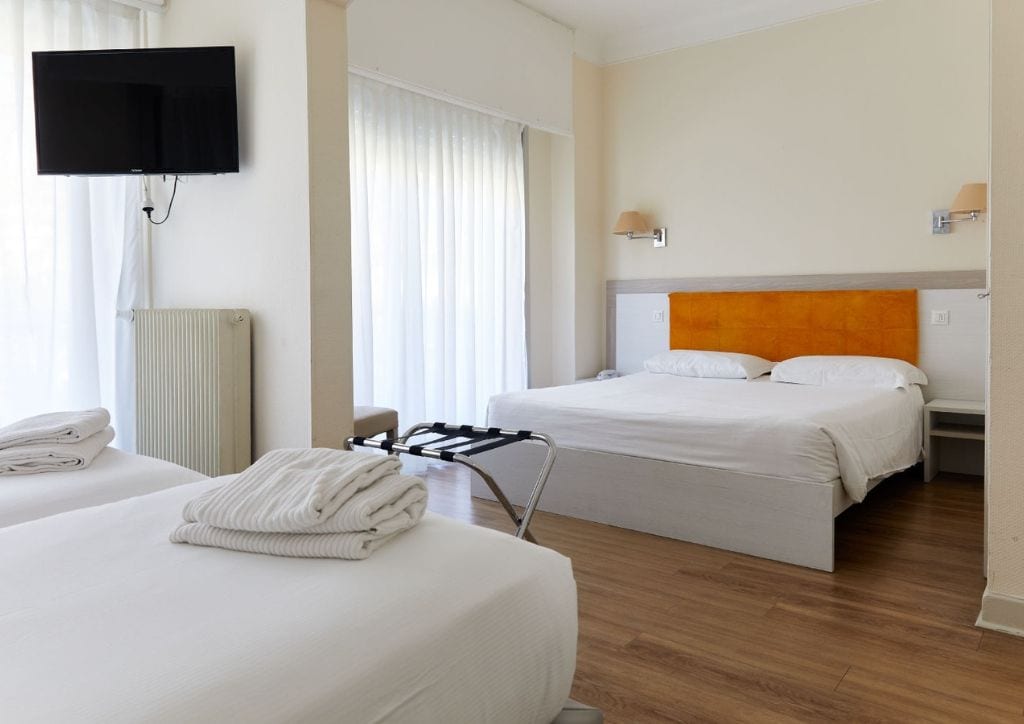 Habitación de hotel con dos camas, un televisor montado en la pared, un radiador y una ventana con cortinas.