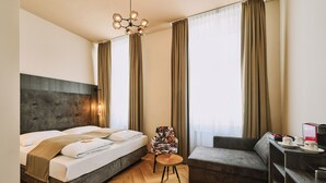 A modern hotel room with a large bed, a sofa, a small round table, and a chair. - Urban Rooms Rathaus - Self Check-In (Vienna)