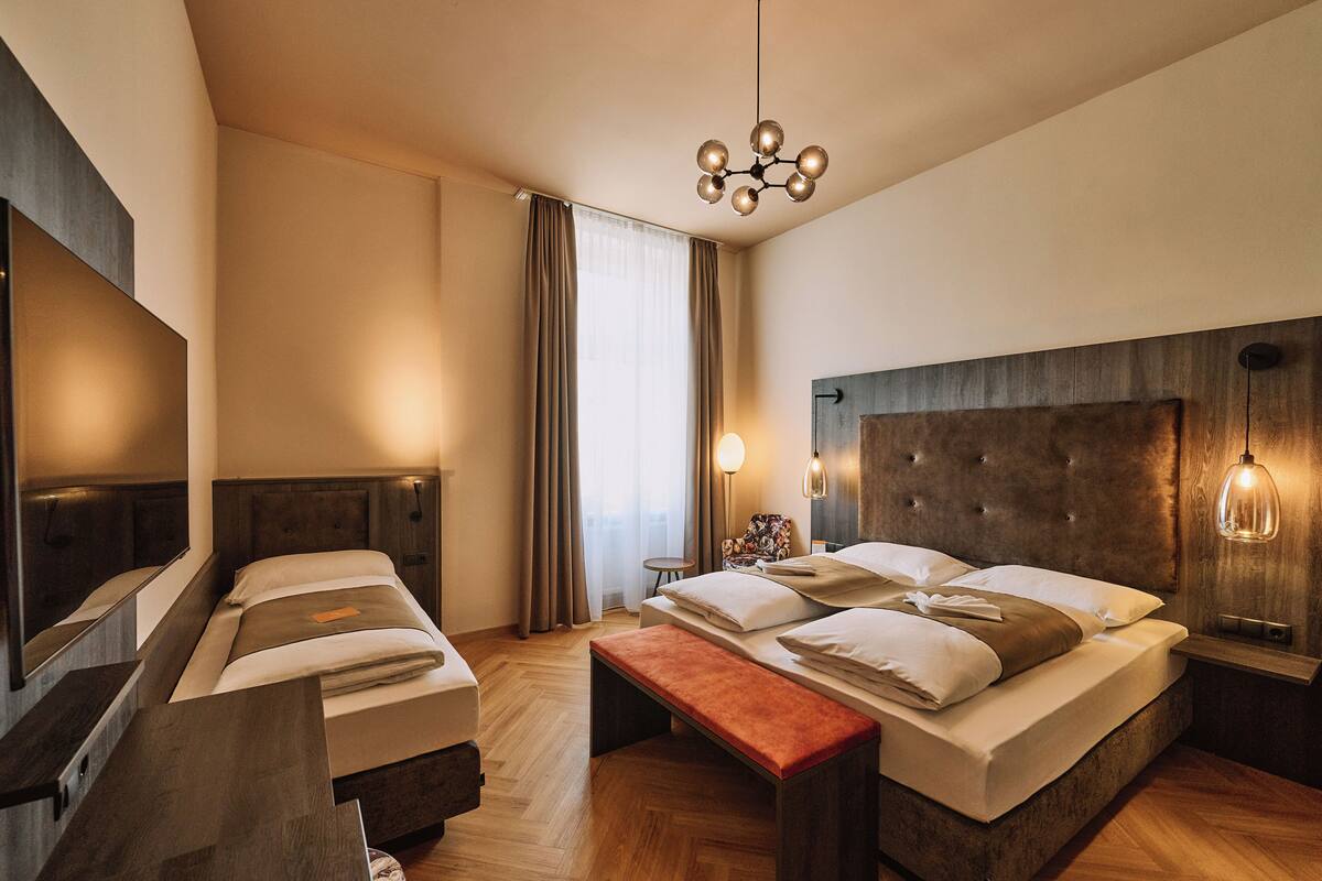 Une chambre d’hôtel moderne avec un grand lit, une banquette, une table de chevet, un miroir et un lustre.