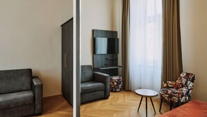 A modern hotel room with a sofa, a chair, a small round table, a TV, and a wardrobe. - Urban Rooms Rathaus - Self Check-In (Vienna)