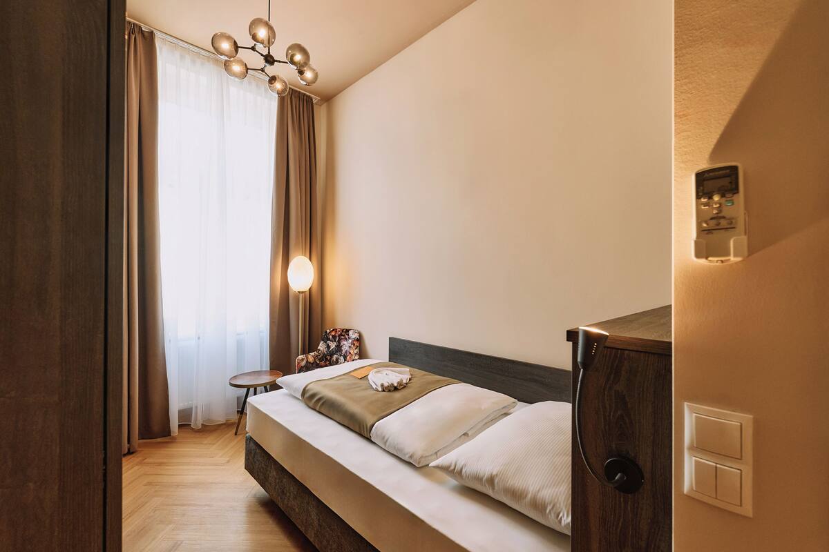 Une chambre d’hôtel moderne avec un plancher en bois, un lit recouvert de draps blancs, une petite table ronde, une chaise et un luminaire fixé au plafond.