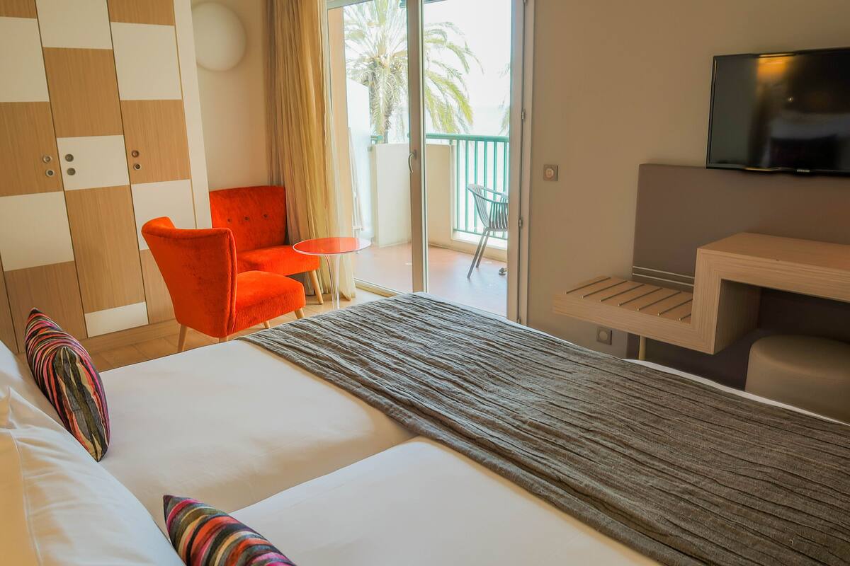 A hotel room with a bed, a small table, a chair, a TV, and a balcony with a view of palm trees.