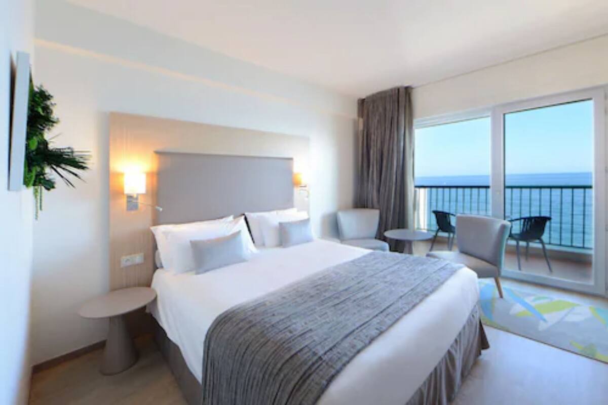 A hotel room with a large bed, a bedside table, a chair, a small round table, and a view of the sea through a balcony.