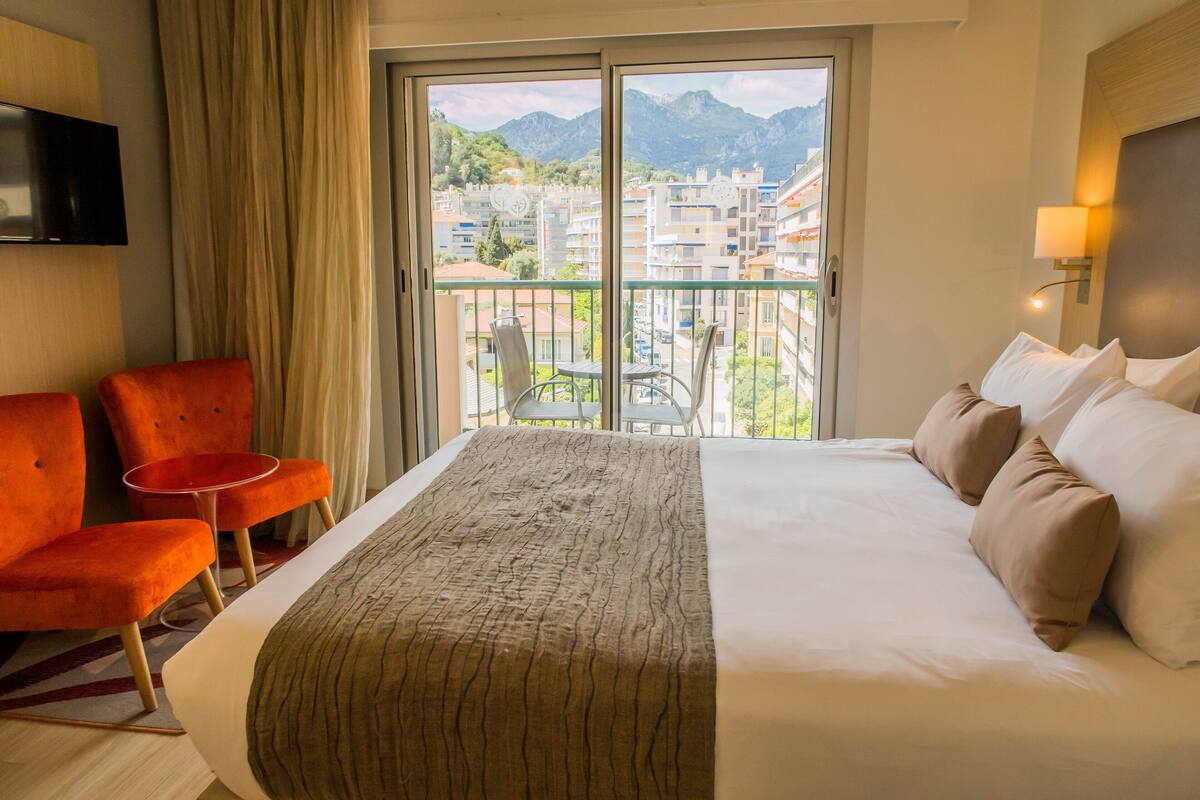 A hotel room with a large bed, two orange chairs, a TV, and a balcony with a mountain view.
