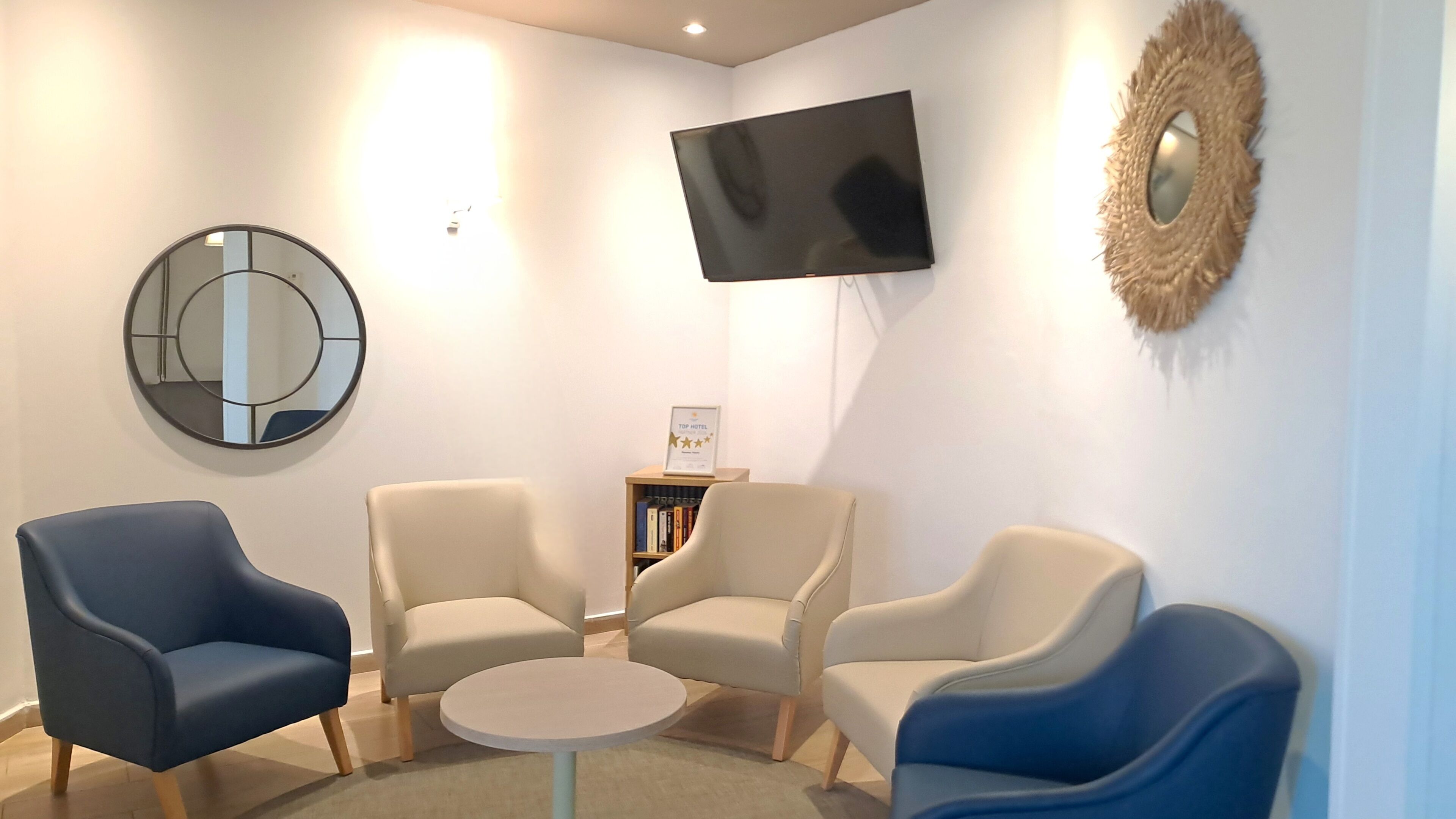 Una habitación con cuatro sillones, una mesita redonda, un televisor, una estantería y un espejo.
