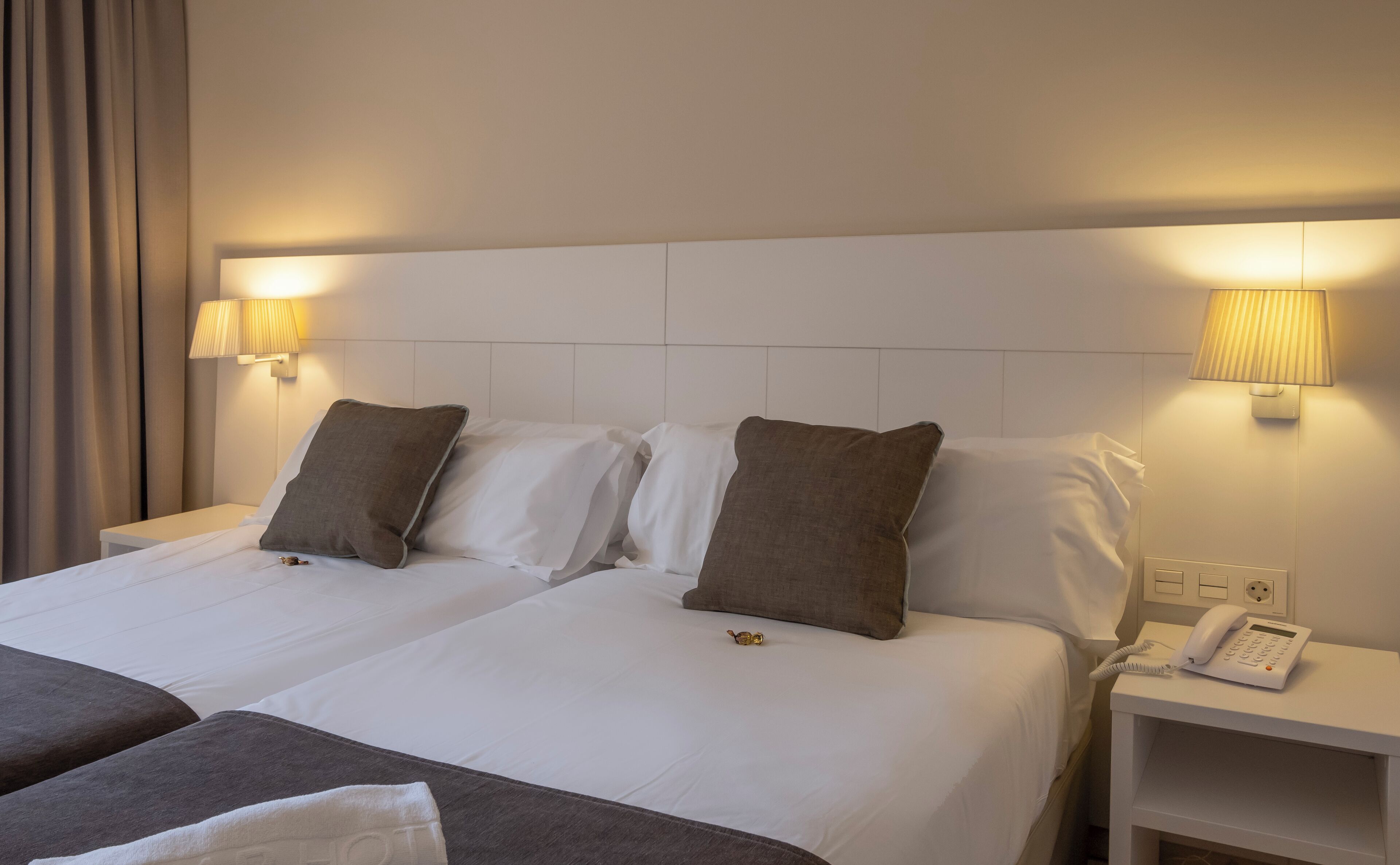 Habitación de hotel con dos camas, cada una con ropa blanca y almohadas marrones. Hay lámparas de noche y un teléfono sobre una mesita.
