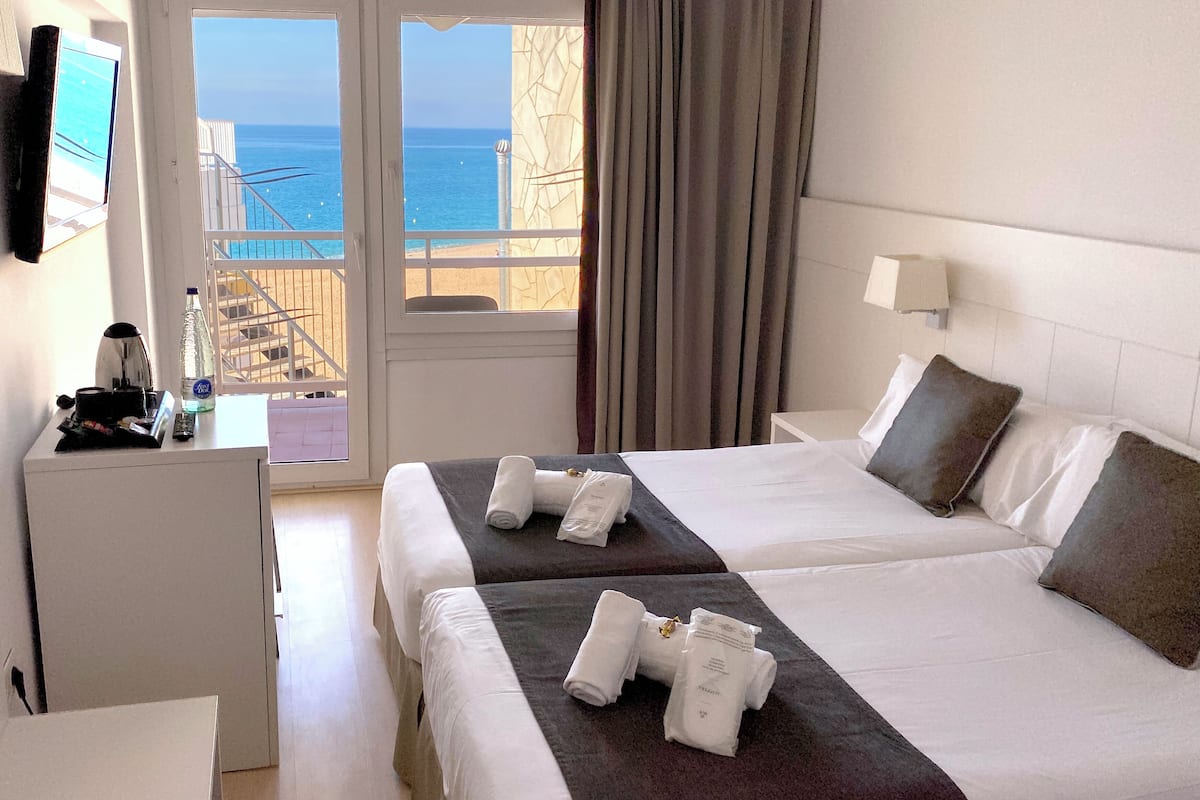 A hotel room with two beds, a desk with a coffee maker, a bottle of water, and a view of the sea.