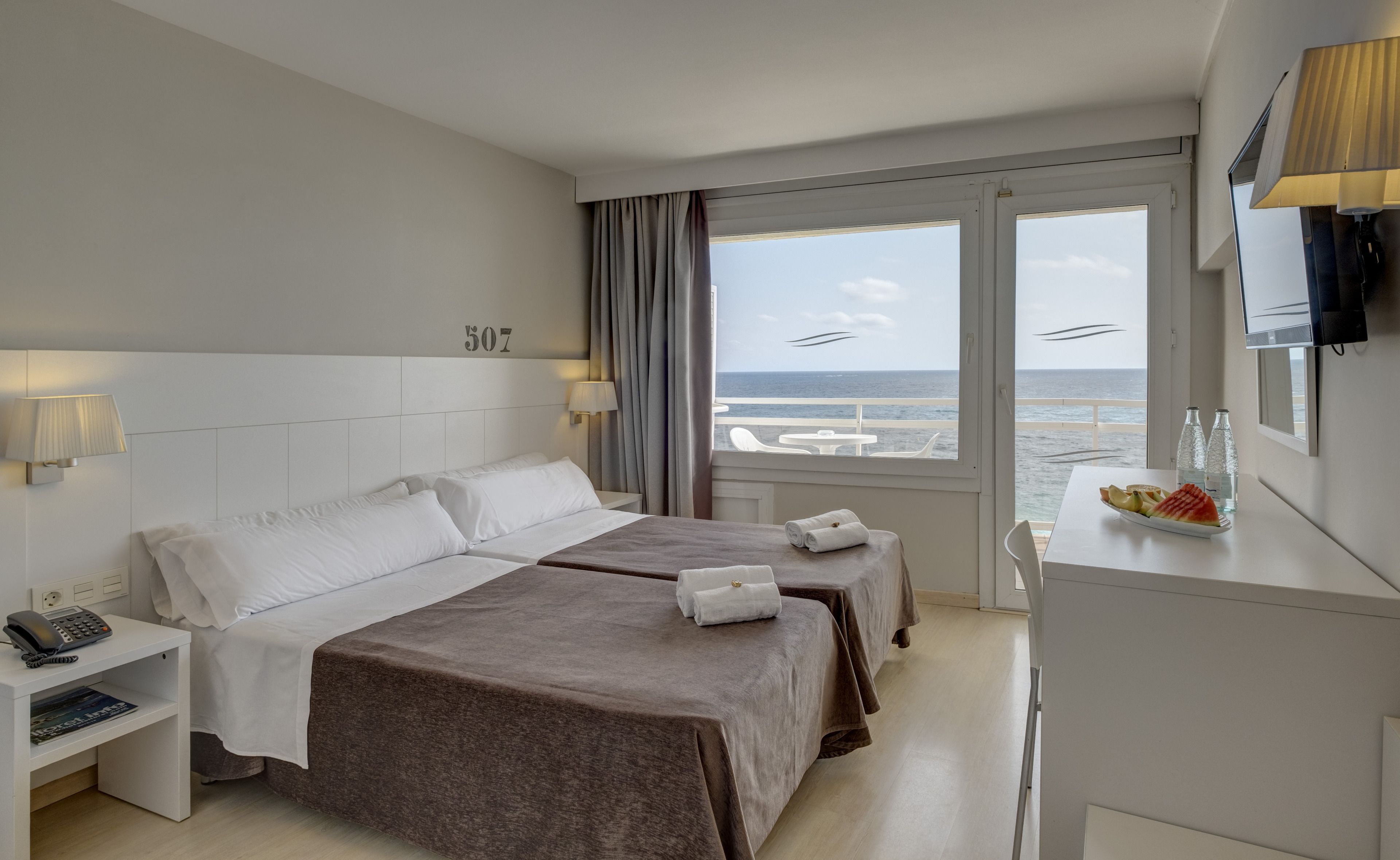 Habitación de hotel con dos camas, un escritorio con lámpara, teléfono y vista al mar.